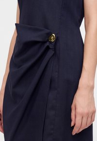 Robe bleu marine avec un tissu drapé rassemblé et fixé par un bouton doré brillant près de la taille, montrant un bras le long du côté.