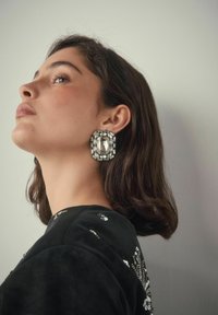 Frau mit kurzem dunklem Haar, die große rechteckige Kristallohrringe und ein schwarzes Oberteil mit Paillettenverzierungen trägt, schaut nach oben.