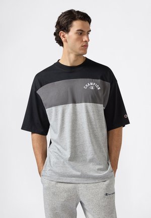 Joven con cabello rizado oscuro que lleva una camiseta Champion negra, gris y gris claro, y pantalones de chándal Champion gris claro, con las manos en los bolsillos.
