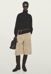 Frau trägt einen schwarzen Rollkragenpullover, einen beigen knielangen Rock, dunkle kniehohe Stiefel, Sonnenbrille und hält eine dunkle Lederhandtasche.