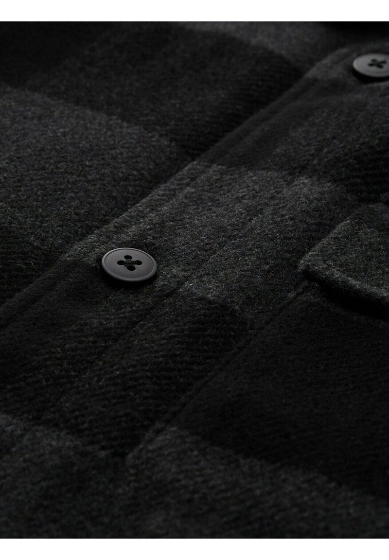 Schwarz- und dunkelgrau kariertes Wollgewebe mit schwarzen Knöpfen und einer Klappentasche an einem Kleidungsstück.