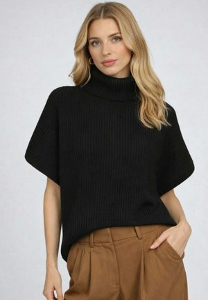Femme blonde portant un pull à col roulé côtelé noir à manches courtes et un pantalon marron taille haute, posant avec une main dans la poche.
