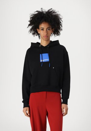 Black cropped hoodie with a blue graphic logo on the chest, featuring a front pocket and drawstring hood, paired with red pants.