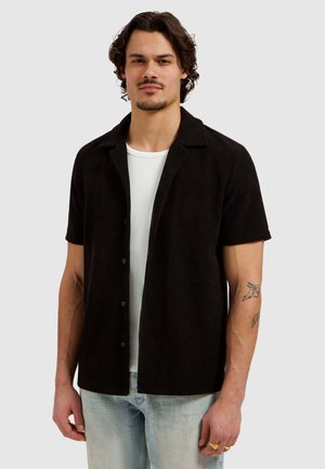 Man met kort krullend haar, gekleed in een zwart overhemd met korte mouwen over een wit T-shirt en lichtblauwe spijkerbroek, staande tegen een effen achtergrond.