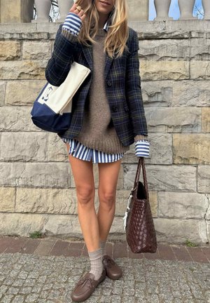 Woman wearing layered outfit with plaid blazer, striped shirt, brown sweater, holding woven brown bag and navy tote, standing by stone wall.