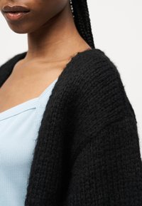 Κοντινό πλάνο ενός ατόμου που φοράει έναν ανοιχτό μπλε ριπ πλεκτό τοπ και μια μαύρη πλεκτή ζακέτα με υφή πάνω από γυμνούς ώμους.