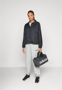 Svart mönstrad jacka, vit t-shirt, grå träningsoverallbyxor med logotyp, svarta sneakers och en svart duffelväska med vit märkning.