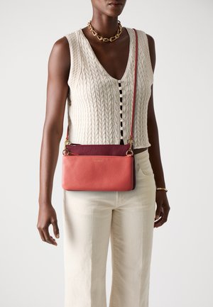 Dark-skinned person wearing a cream cable knit sleeveless vest, cream pants, gold chain necklace, and a two-tone red crossbody bag.