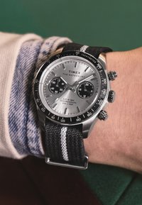 Armbåndsur med sort og hvidt nylonstrap, sølv chronograph urskive, tachymeter ramme, båret på en persons håndled over en beige og blå skjorteærme.