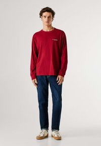 Maglietta rossa a maniche lunghe con collo rotondo, abbinata a jeans blu scuro e sneakers. La maglietta presenta un piccolo logo stampato sul petto.