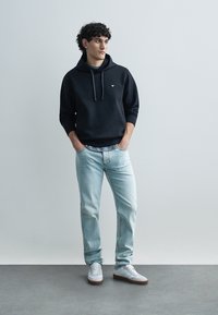 Μαύρο φούτερ με κουκούλα και κορδόνια, ανοιχτό μπλε τζιν και λευκά αθλητικά παπούτσια. Σημαντικές λεπτομέρειες περιλαμβάνουν ένα μικρό λογότυπο στο φούτερ και άνετη εφαρμογή.