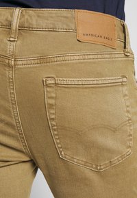 Khakifärgad denimjeans med synlig bakficka och rundad sömdetalj. Midjan har en brun läderetikett med "AMERICAN EAGLE" präglat.