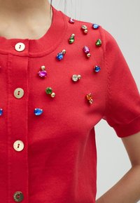 Cardigan en tricot rouge avec un col rond et des manches courtes, orné de bijoux multicolores et de boutons dorés.