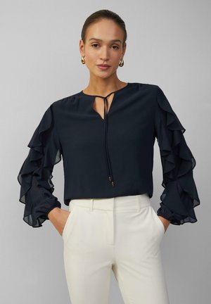 Blouse marine avec des manches en dentelle à volants, dotée d'un décolleté en forme de clé et d'un détail à nouer. Associée à un pantalon ajusté de couleur claire.