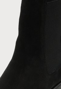 Gros plan d'une bottine en daim noir avec panneau latéral élastique et couture visible sur un fond clair.