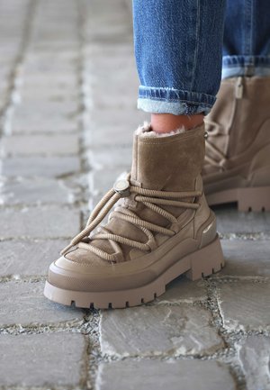 Tan enkellaarsjes van suede met een pluche voering, voorzien van dikke, gestructureerde zolen en veterlussen. Het ontwerp bevat een metallic accent.