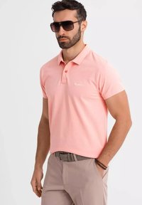 Rosa bomullspolo med klassisk krage, tvåknapplist och liten vit logotyp på bröstet; kombinerad med beige byxor.