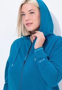 Vaaleatukkainen nainen, joka käyttää sinistä vetoketjullista hupparia, jossa huppu on osittain päällä ja hän pitää hupparin nyöriä lähellä kasvojaan, valkoista taustaa vasten.