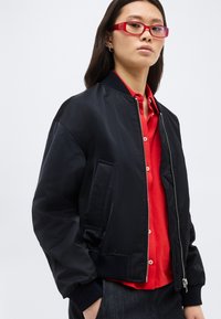 Νεαρή γυναίκα με μακριά σκούρα μαλλιά, που φοράει κόκκινα γυαλιά, μαύρο μπουφάν τύπου bomber και κόκκινο πουκάμισο με κουμπιά, στέκεται μπροστά σε απλό φόντο.