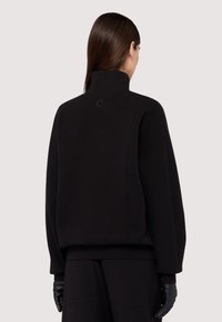 Černá mikina s vysokým límcem a volným střihem, vyrobená z měkké tkaniny. Obsahuje embosované logo na zadní straně a boční kapsy.