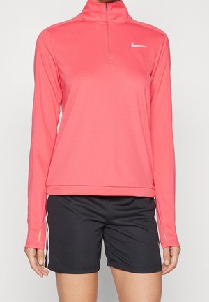 Kobieta ubrana w koralowo-różowy długi rękaw Nike z suwakiem oraz czarne sportowe szorty z białymi paskami po bokach, stoi na jednolitym tle.