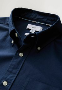 Chemise bleu marine à boutonnière avec boutons blancs et étiquette de col indiquant "NEXT", taille moyenne, fabriquée au Bangladesh, 100 % coton.