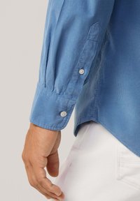 Lichtblauwe corduroy blouse met lange mouwen, knoopmanchetten en een licht gebogen zoom. Hand en witte broek gedeeltelijk zichtbaar.