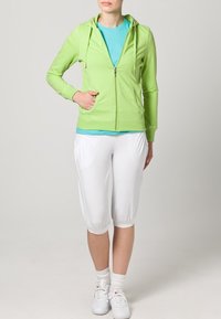 Ljusgrön zip-up hoodie med dragsko, bärs över en turkos tröja. Vita knälånga shorts, vita sneakers och ankelsockor.