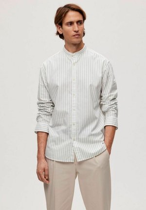 Uomo in camicia bianca a maniche lunghe con sottili righe verticali e pantaloni beige, in piedi con una mano in tasca su uno sfondo semplice.