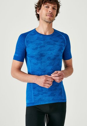 Uomo che indossa una maglietta sportiva blu aderente a maniche corte con un motivo sottile, in piedi davanti a uno sfondo chiaro uniforme, con le mani intrecciate.