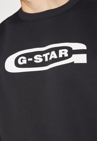 Černé tričko s kulatým výstřihem a výrazným bílým logem G-STAR uprostřed na hrudi.