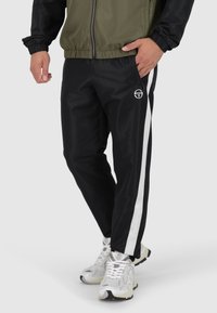 Schwarze Jogginghose mit weißen Seitenstreifen, aus glattem Stoff gefertigt. Hat eine enganliegende Taille und ein Logo auf der linken Hüfte. Getragen mit weißen Sneakers.