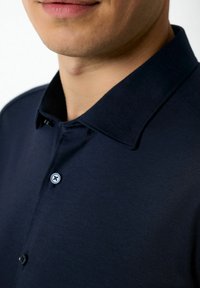 Marineblaues Poloshirt aus strukturiertem Material, mit einem spitzen Kragen, zwei Knöpfen und kontrastierenden Knopfdetails.