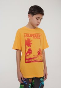 T-shirt di cotone gialla con una grafica rossa di palme e un surfista, con il testo "SUNSET CALIFORNIA" sopra, vestibilità rilassata, maniche corte.