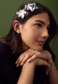 Silberne Haarklammern mit Pailletten in Sternform, bestehend aus zwei Sternen, befestigt mit einem Metallverschluss. Die Haare sind dunkel und glatt, vor neutralem Hintergrund.
