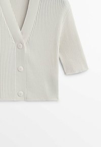 Cardigan en maille côtelée couleur crème avec trois boutons et manches trois-quarts sur fond blanc.