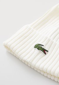 White ribbed beanie made from knit material, featuring a green crocodile logo embroidered on the folded edge.