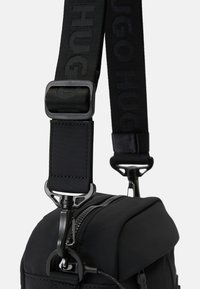 Mala de slings em tecido preto com uma alça texturizada com marcação em relevo, hardware de clip de metal e detalhe visível na zipper da mala.