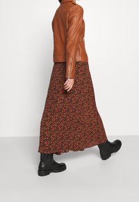Giacca di pelle marrone con dettagli in zip, gonna maxi nera a fantasia floreale in tonalità di rosa e giallo, e stivali neri chunky.