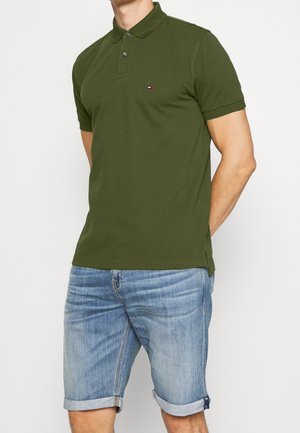 Mężczyzna w oliwkowozielonej koszulce polo z małym logo i jasnoniebieskich podwiniętych szortach dżinsowych na gładkim tle.