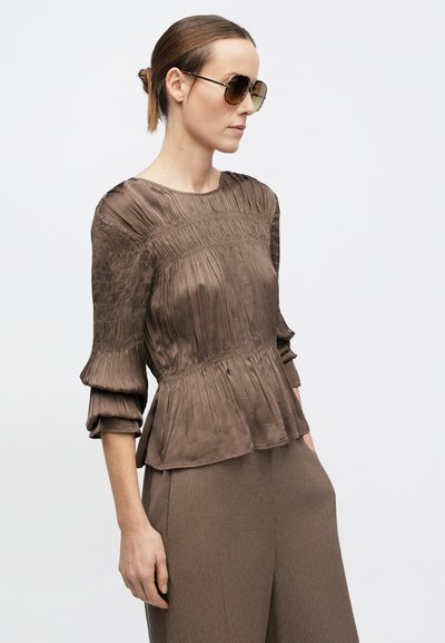 Barna, strukturált peplum felső pliszírozott mintával, háromnegyedes ujjakkal és kerek nyakkivágással, hozzáillő nadrággal és napszemüveggel párosítva.