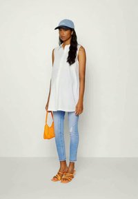 Camisola branca sem mangas, boné azul claro, jeans azul claro, sandálias de salto laranja e uma pequena bolsa laranja.