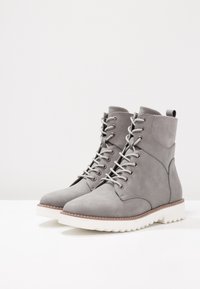 Bottes cheville en daim gris avec bout arrondi, design à lacets et œillets argentés. Semelle épaisse en caoutchouc blanc avec grip texturé et accents marron.