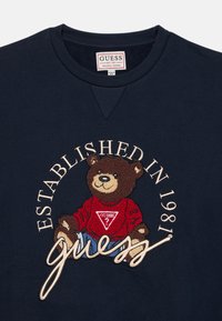 Sweatshirt marine avec un graphique rond représentant un ours en chemise rouge, texte brodé "ÉTABLI EN 1981" et "guess". Tissu au toucher doux.