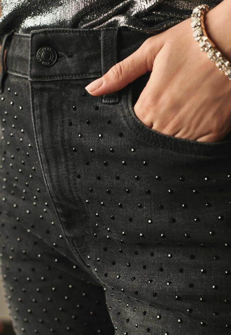 Jean en denim noir avec une surface texturée ornée de petits strass. Gros plan sur la zone de la poche avec un détail de bouton.