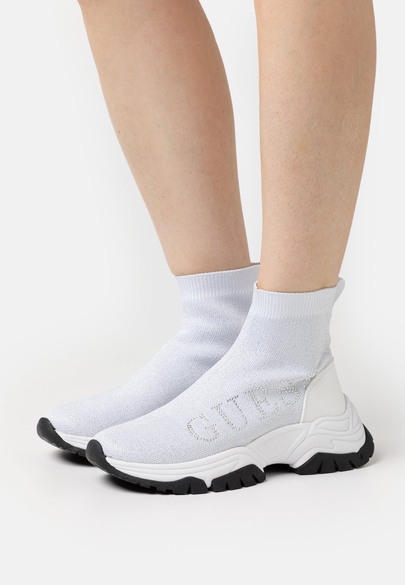 Baskets chaussettes blanches avec semelles noires et logo «GUESS» en strass, portées sur jambes nues sur un fond uni.