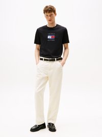 T-shirt noire à manches courtes avec un logo coloré "Tommy Hilfiger", associé à un pantalon droit couleur crème et des chaussures noires brillantes.