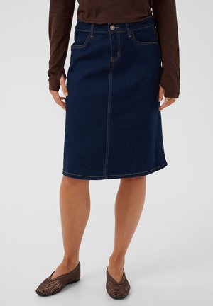 Kvinde iført en mørkeblå denim nederdel til knæet, brun langærmet top og mørke vævede flade sko, stående med afslappede hænder.