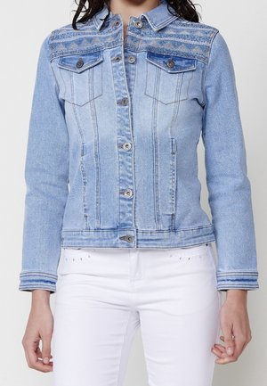 Lichtblauwe spijkerjas met een patroon op het schouderstuk, knoopsluiting voor, borstzakken en lange mouwen, gedragen met een witte broek.