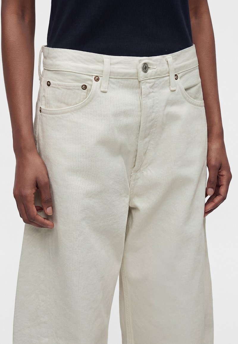 Personne portant un pantalon large taille haute écru avec des boutons métalliques et des rivets, associé à un haut foncé rentré.